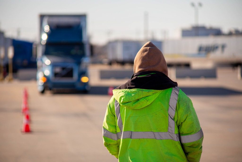 CDL instructor in a high-visibility safety vest spotting a student driver practicing backing maneuvers on a semi-truck cone course