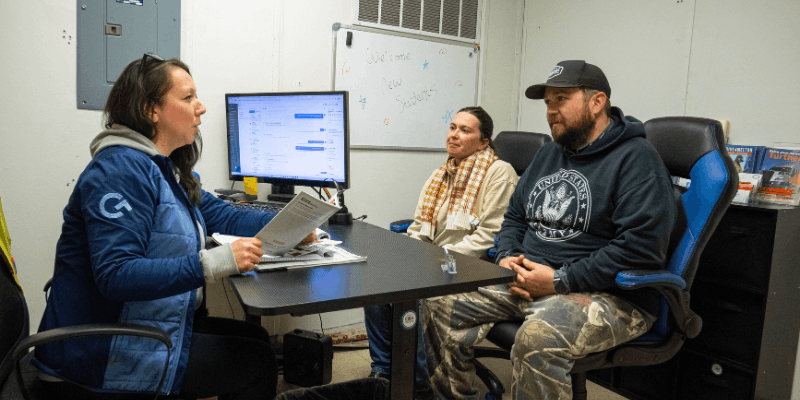 Admissions advisor holding papers and speaking with two potential students at desk